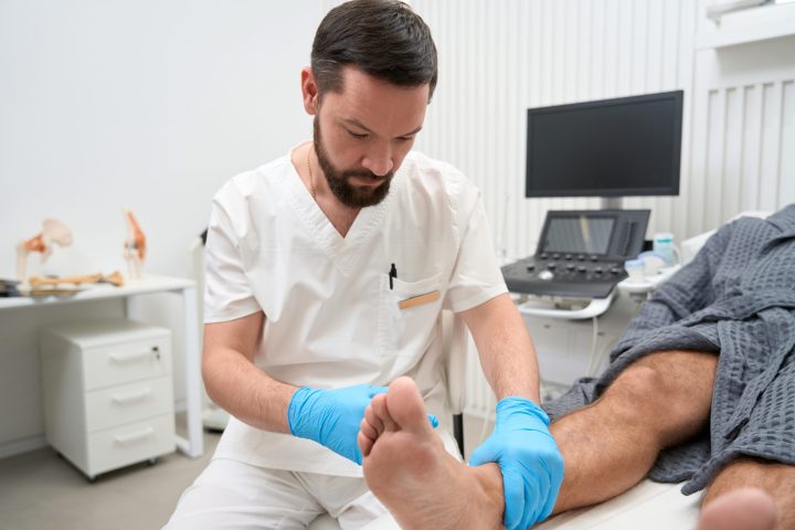Focused brunet doctor in surgical scrubs and gloves examining ankle of a patient in hospital bed